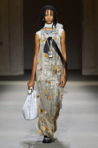 Model on a runway wearing a sheer champagne gown with gold stars, layered necklaces, and a white handbag with gold hardware; she also has a black scarf tie draped over the dress.