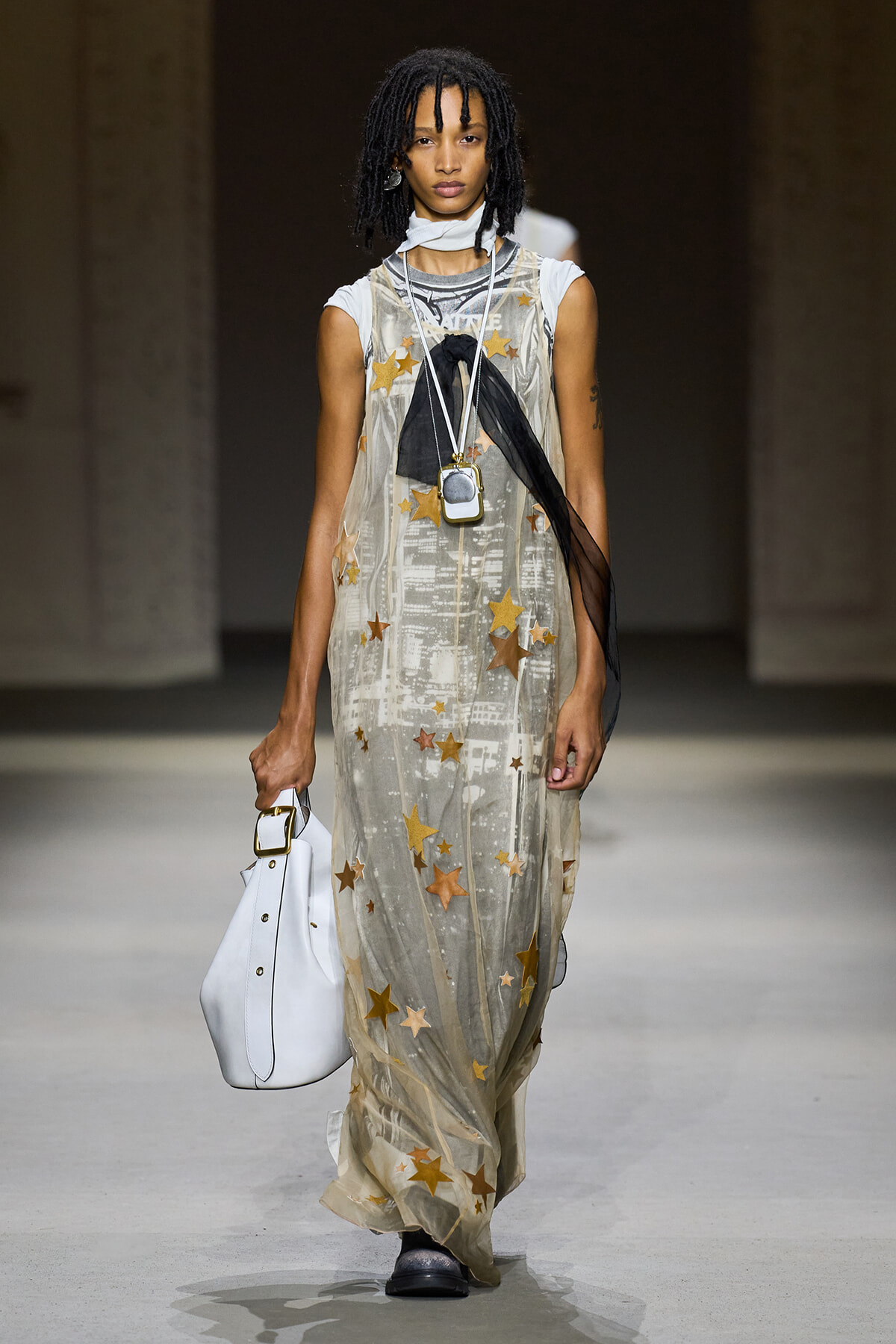 Model on a runway wearing a sheer champagne gown with gold stars, layered necklaces, and a white handbag with gold hardware; she also has a black scarf tie draped over the dress.