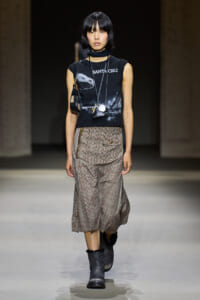 Runway model wearing a black sleeveless graphic tee, layered necklaces, a plaid midi skirt with zippers, and gray fur-trim boots.