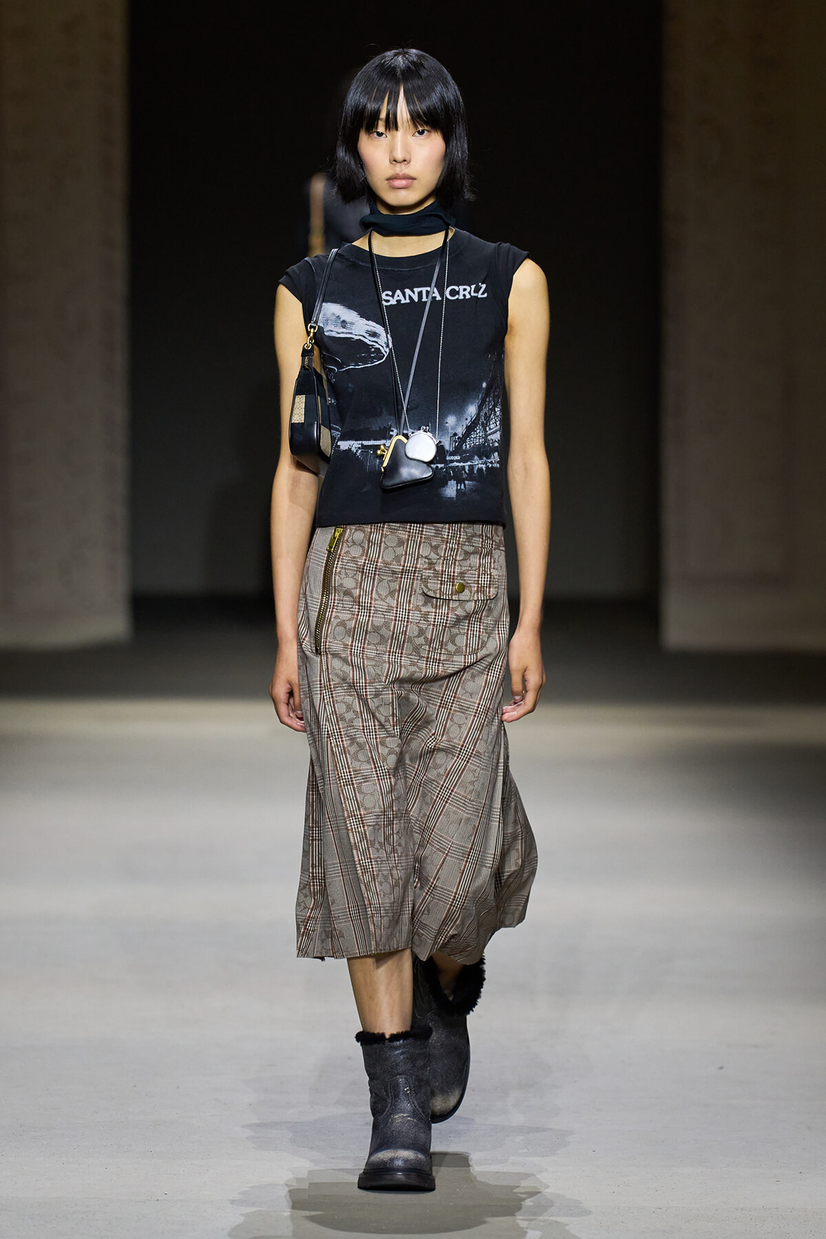 Runway model wearing a black sleeveless graphic tee, layered necklaces, a plaid midi skirt with zippers, and gray fur-trim boots.