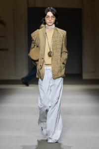 Model on a runway wearing a beige patterned blazer over a tan turtleneck, light-washed wide-leg jeans, round glasses, and a tan circular bag crossbody accessory.
