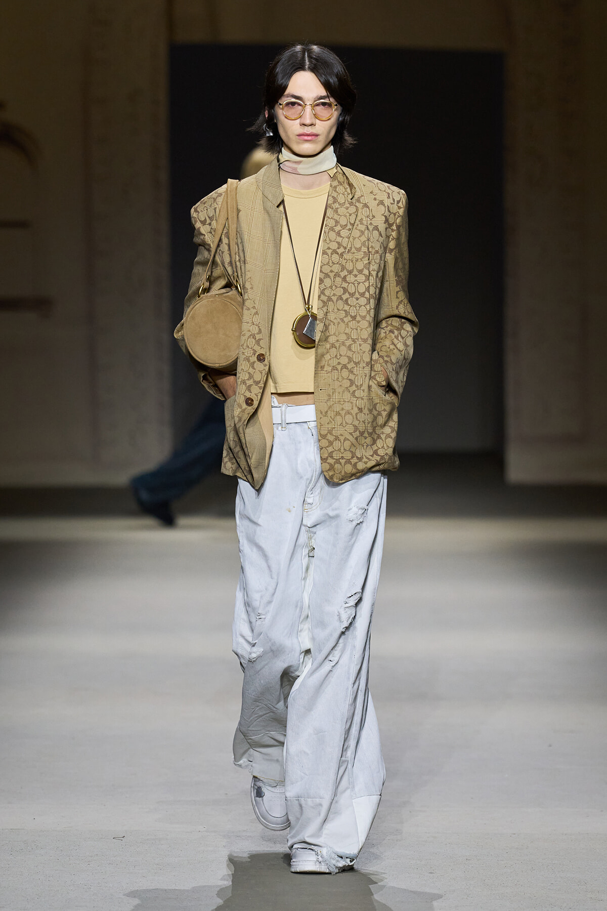 Model on a runway wearing a beige patterned blazer over a tan turtleneck, light-washed wide-leg jeans, round glasses, and a tan circular bag crossbody accessory.