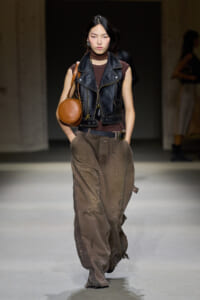 Model walking a runway in a sleeveless black leather vest over a brown top, with oversized distressed brown cargo pants and a round tan shoulder bag.
