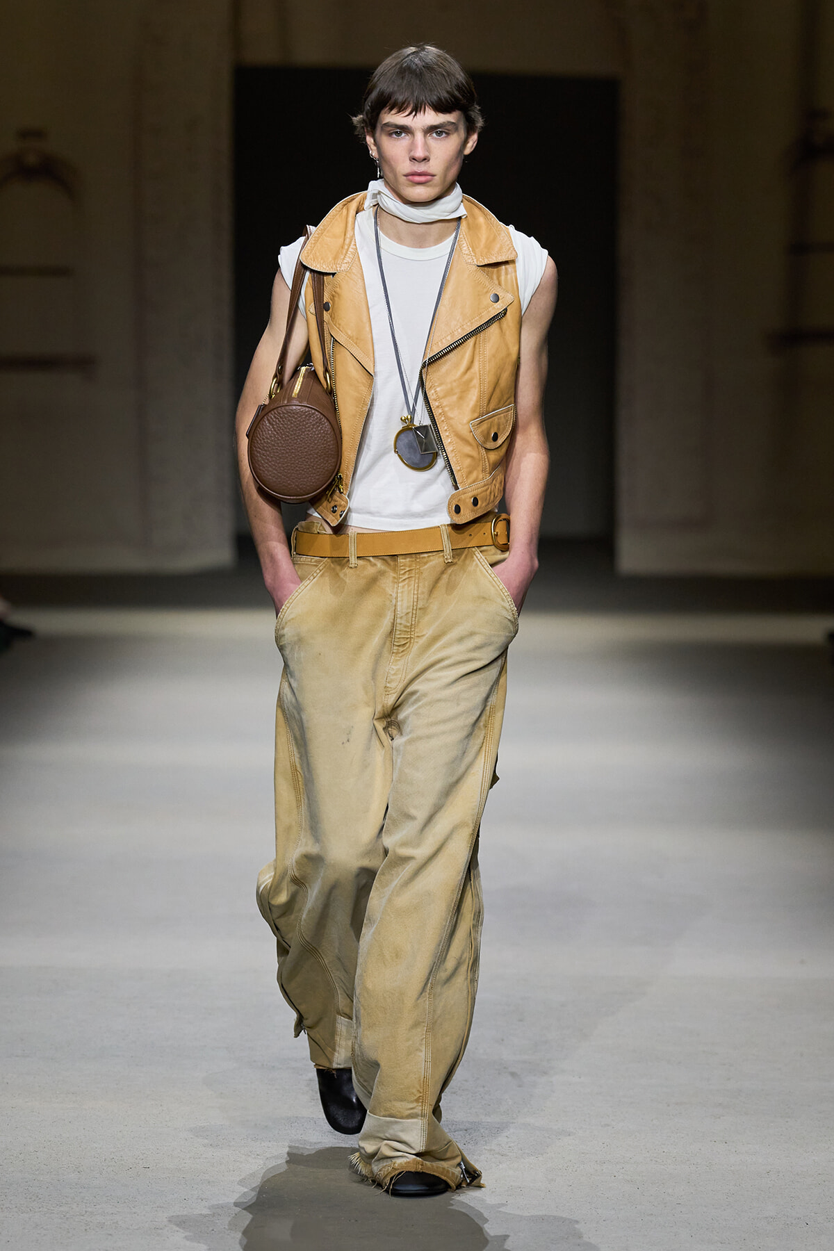 Model on runway wearing a tan leather vest over a white top, beige baggy pants, brown round crossbody bag, and layered necklaces with a pendant.