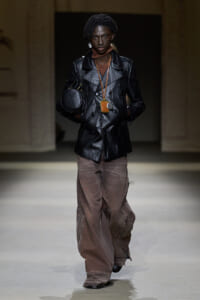 Model on a runway wearing a black leather double-breasted jacket, brown cargo pants, a small round black handbag, and layered necklaces with pendants.