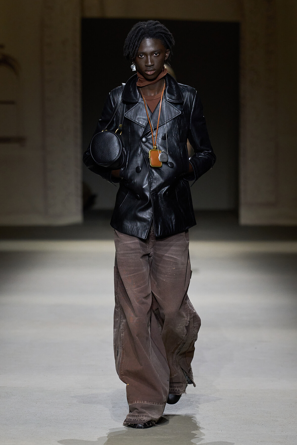 Model on a runway wearing a black leather double-breasted jacket, brown cargo pants, a small round black handbag, and layered necklaces with pendants.