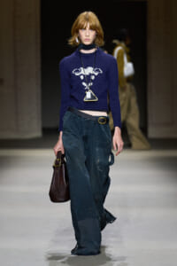 Model walking a fashion runway in a navy cropped sweater with a white crest, dark baggy jeans, and a brown handbag.