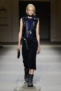 Model walking runway in a black sleeveless leather vest over a navy top, paired with a black zippered skirt and gray ankle boots, accessorized with long necklaces and a small round handbag.