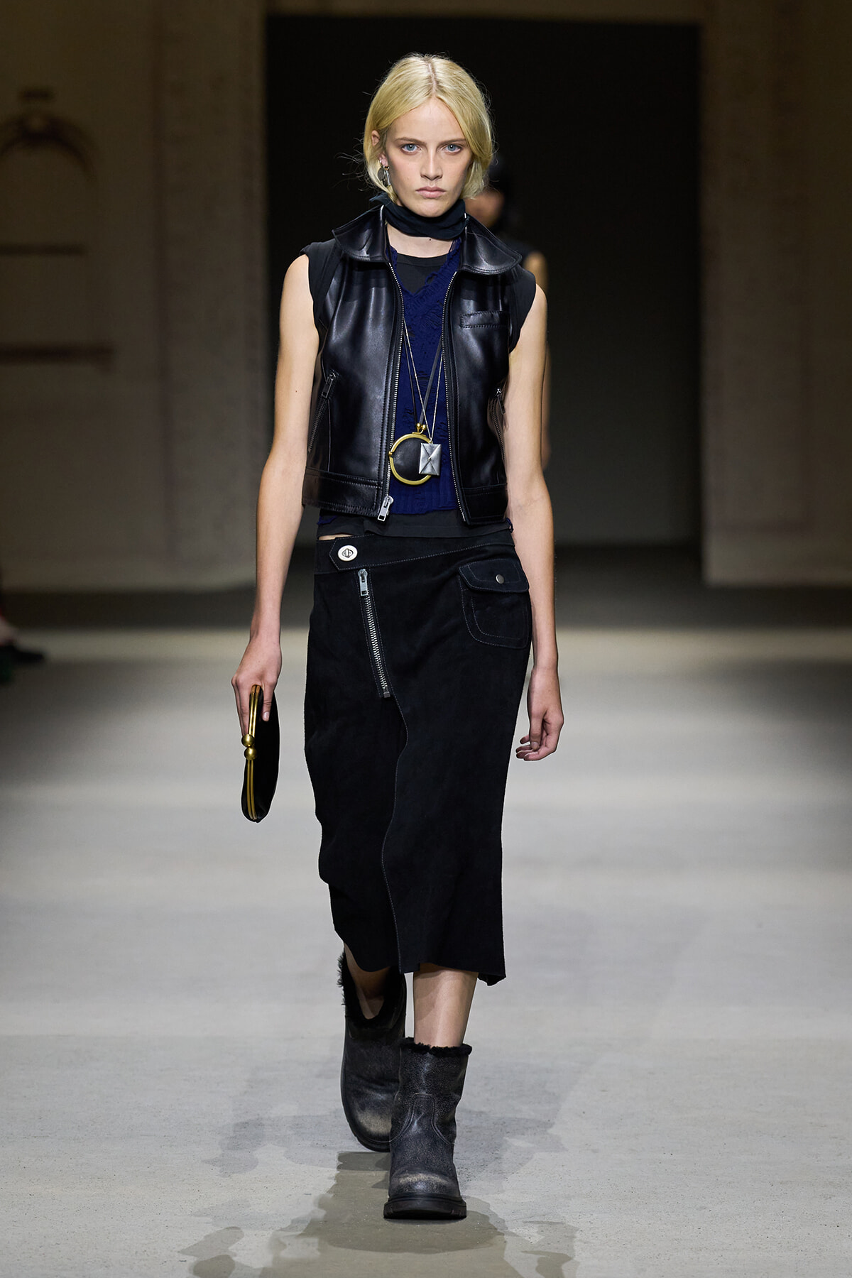 Model walking runway in a black sleeveless leather vest over a navy top, paired with a black zippered skirt and gray ankle boots, accessorized with long necklaces and a small round handbag.