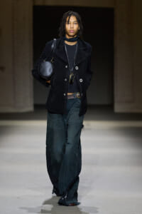 Model on a runway wearing a black suede double-breasted coat, black top, and oversized dark pants, carrying a round black handbag and layered necklaces.