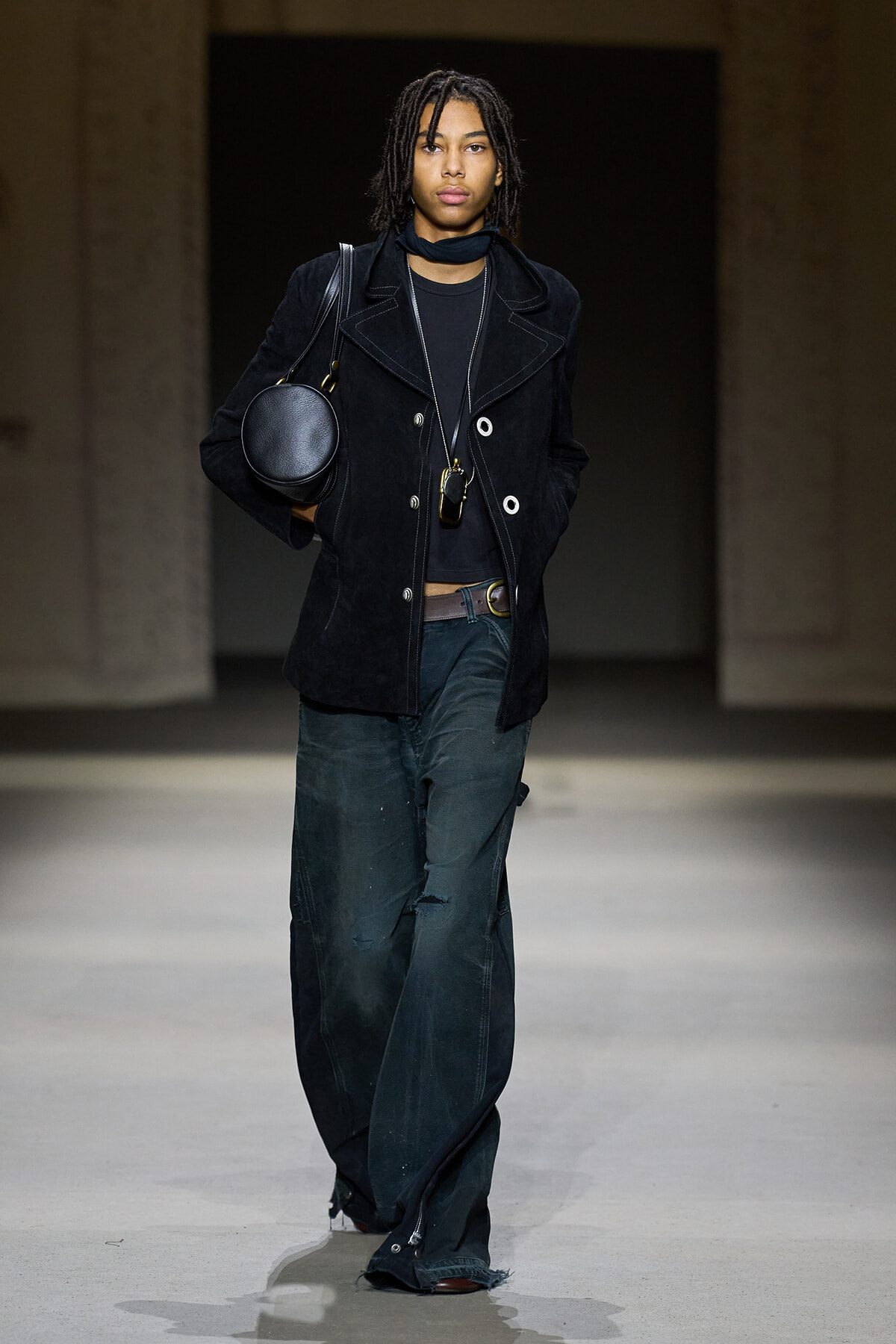 Model on a runway wearing a black suede double-breasted coat, black top, and oversized dark pants, carrying a round black handbag and layered necklaces.