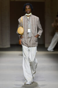 Model walking a runway in an oversized plaid blazer with white paint-splattered jeans, carrying a round yellow bag over the shoulder and wearing yellow sunglasses with a neck scarf and badge.