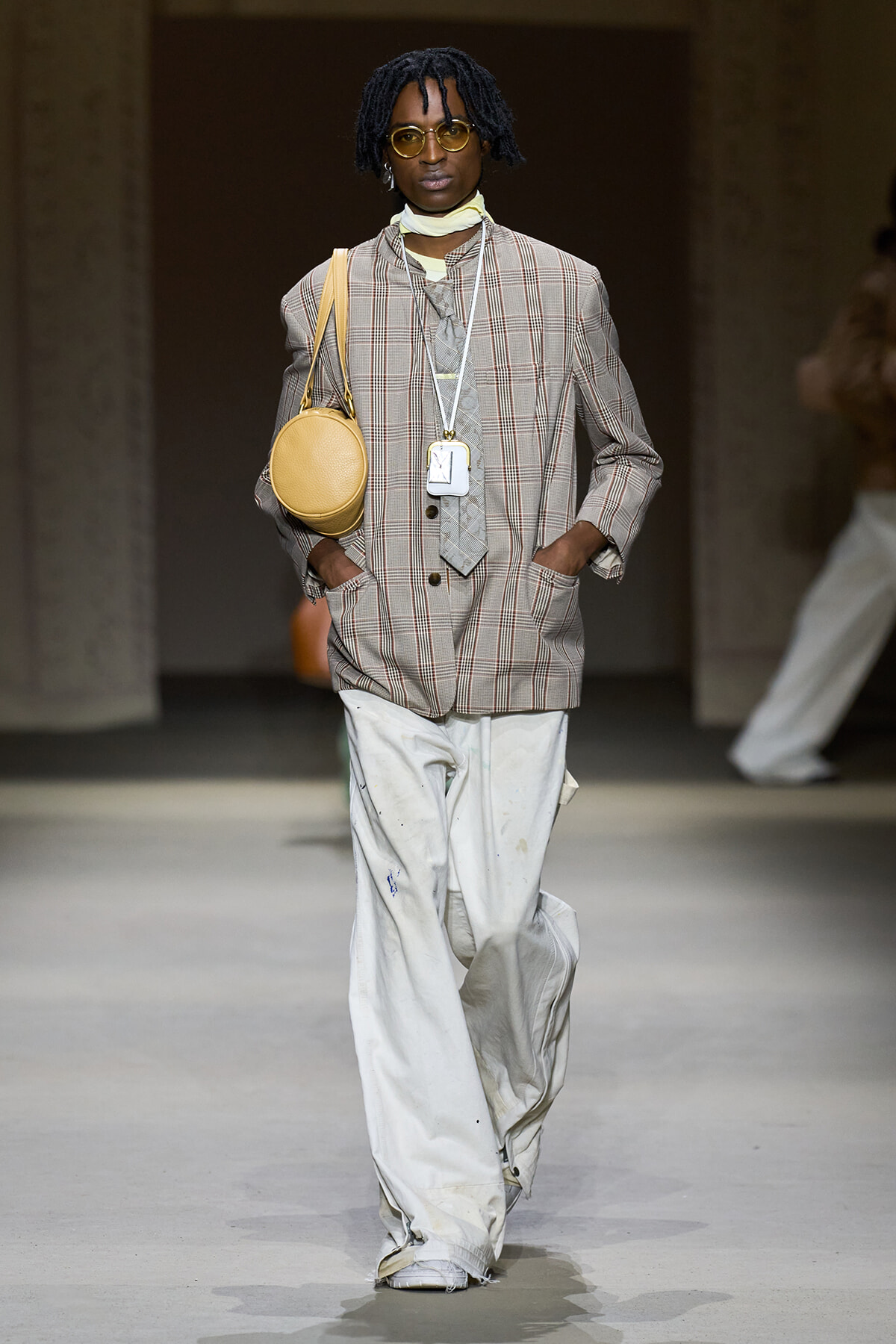 Model walking a runway in an oversized plaid blazer with white paint-splattered jeans, carrying a round yellow bag over the shoulder and wearing yellow sunglasses with a neck scarf and badge.