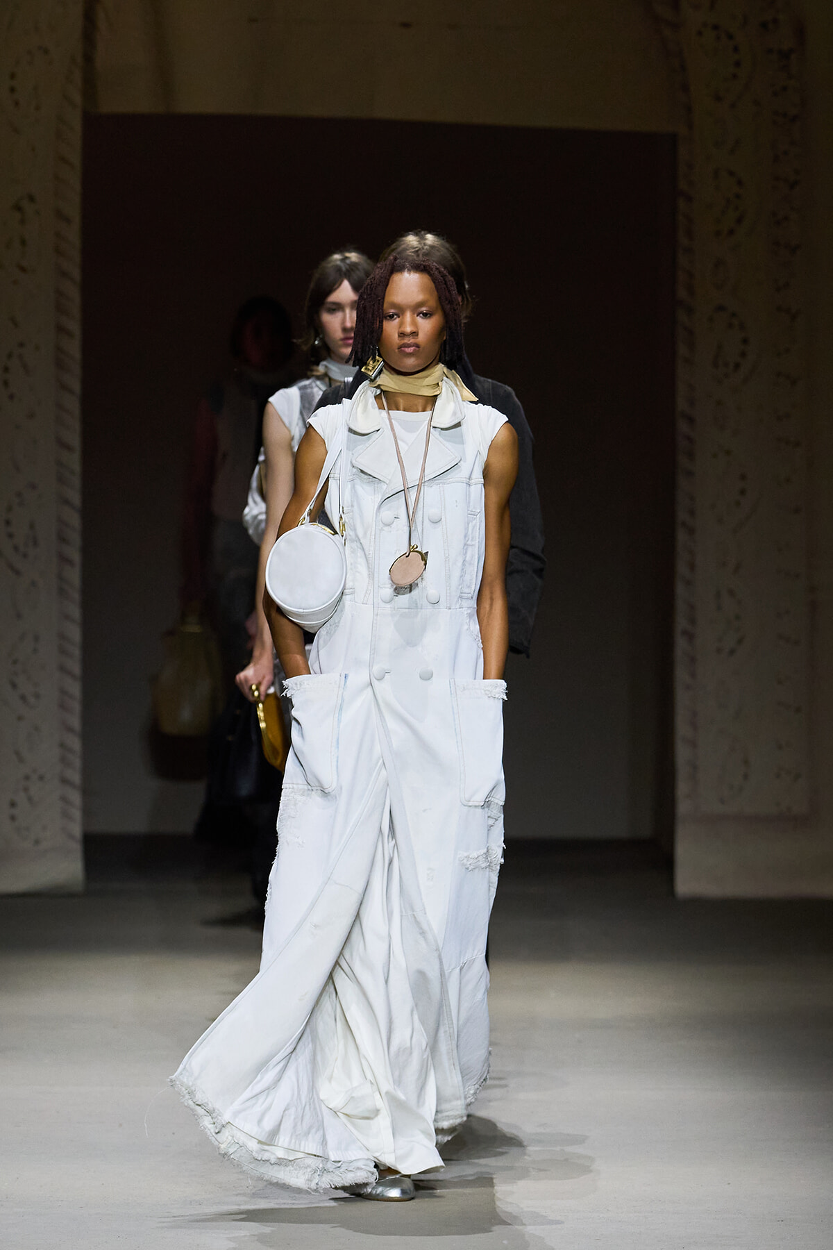 Model walks the runway in an oversized white denim utility jumpsuit with large pockets, layered necklaces, and a round white purse.