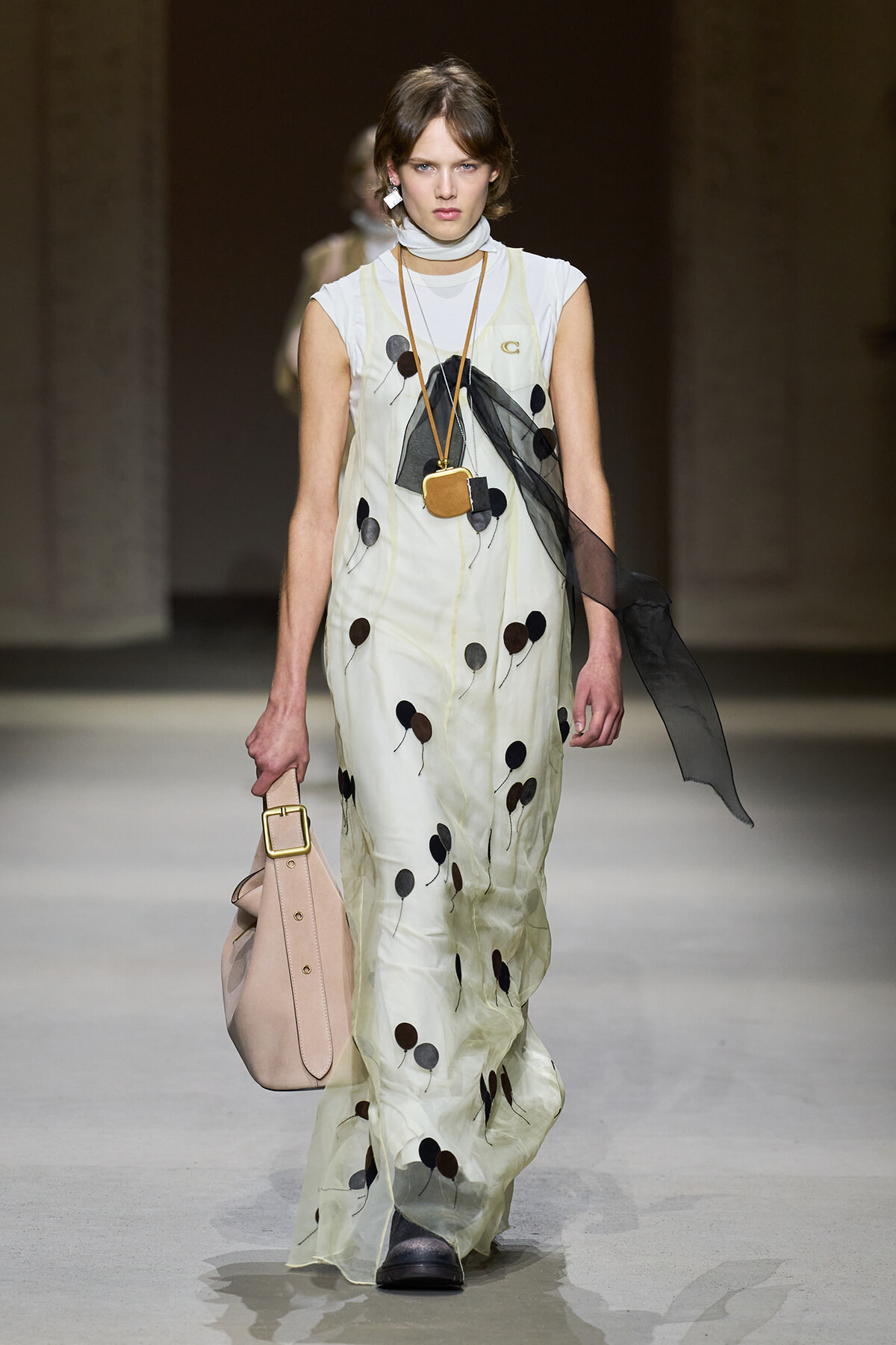Model walks the runway in a pale cream sleeveless maxi dress with black balloon-like spots, accessorized with a long pendant necklace and a sheer black scarf.