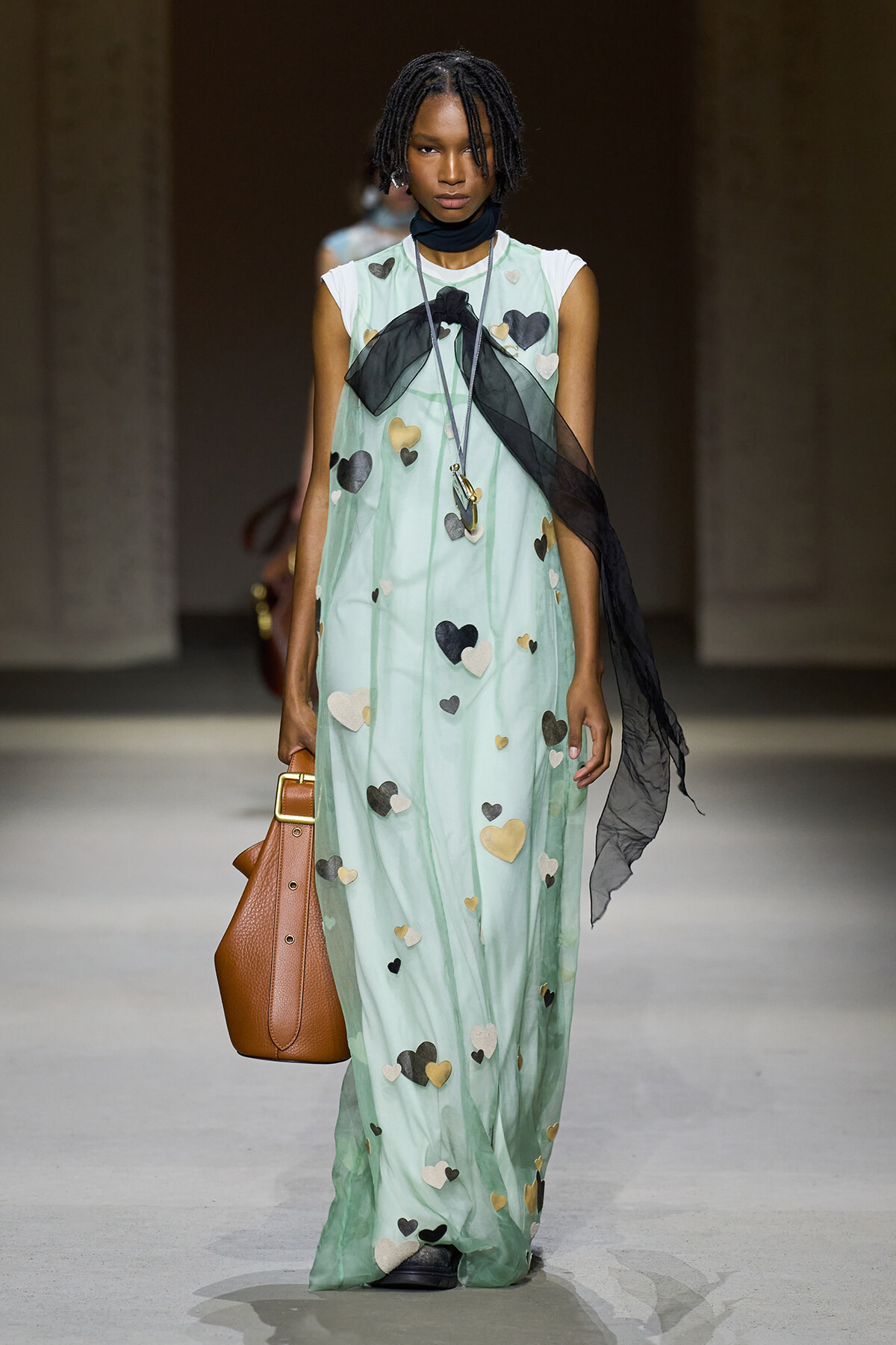 Model walks a runway in a mint-green maxi dress covered with pastel heart appliqués, paired with a sheer black scarf and brown leather handbag.