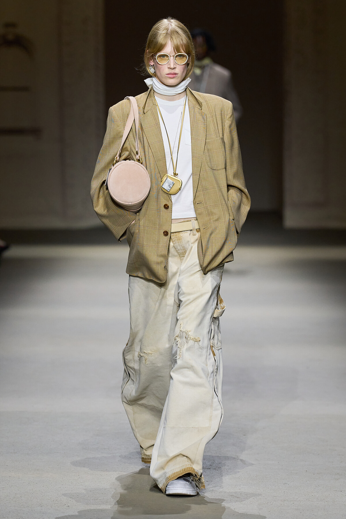 Model walking runway in oversized beige plaid blazer, white top, baggy distressed cream pants, round glasses, and pale pink circular handbag.