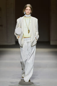 Model walking a fashion runway in a pale beige suit with oversized pants, mint top, and accessorized with a round white bag and long gold necklaces.