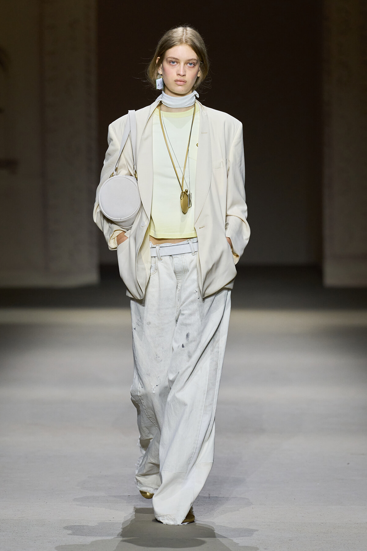 Model walking a fashion runway in a pale beige suit with oversized pants, mint top, and accessorized with a round white bag and long gold necklaces.