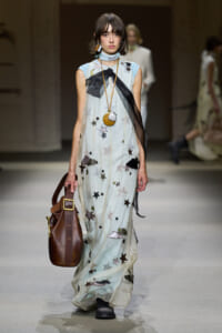 Model wearing a sheer, star-and-cloud printed dress on a runway, carrying a brown leather bag and wearing bold jewelry.