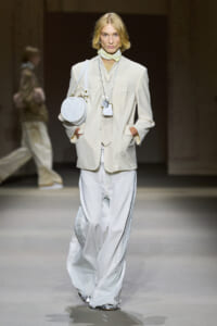 Model on a runway wearing a light cream suit with an oversized blazer and wide white pants, accessorized with a round white shoulder bag and layered necklaces.