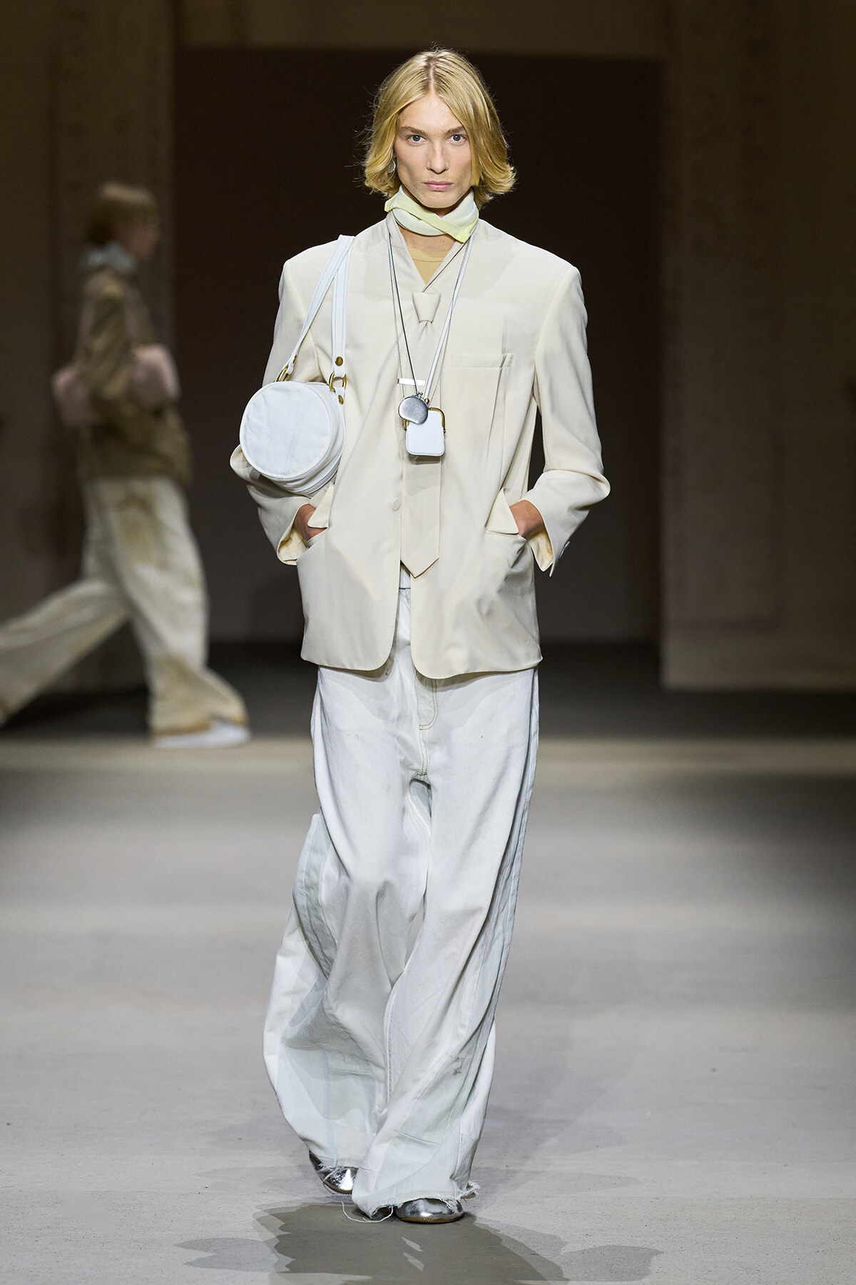 Model on a runway wearing a light cream suit with an oversized blazer and wide white pants, accessorized with a round white shoulder bag and layered necklaces.