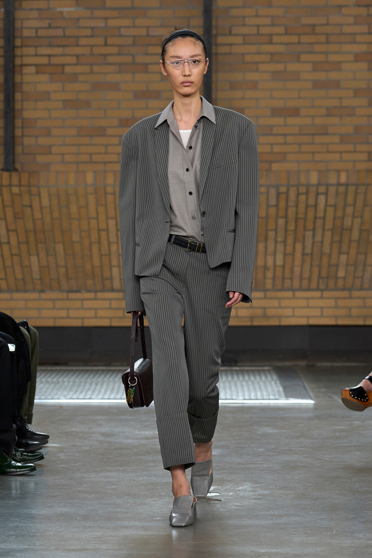 Model wearing a gray pinstripe suit with vest, light blouse, and silver loafers on a fashion runway, carrying a brown handbag.