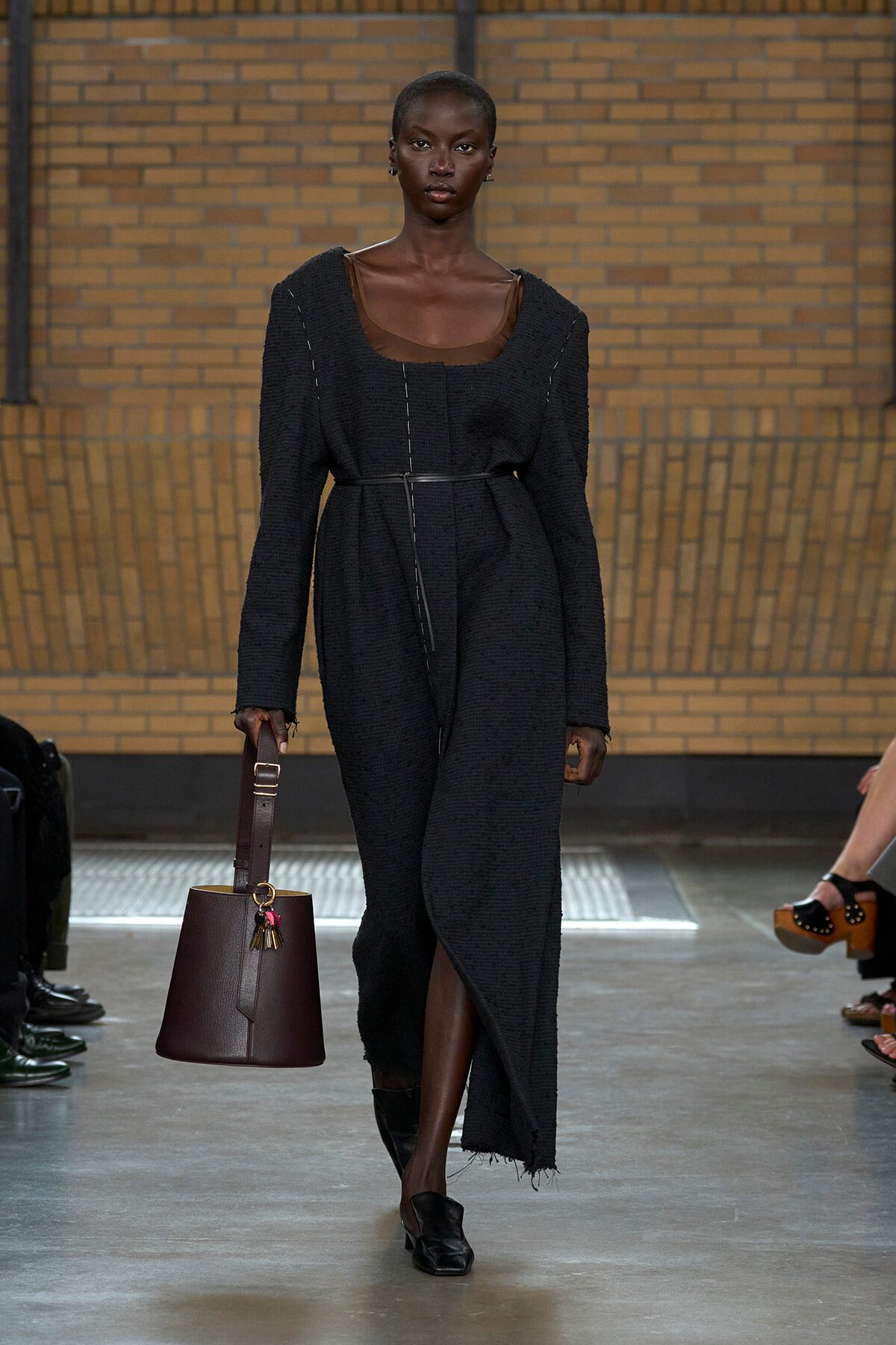 Model walking a fashion runway in a black textured long coat with a belt, holding a burgundy handbag.