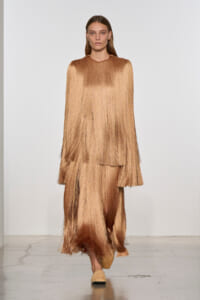 Model walking the runway in a metallic bronze fringe dress with layered, floor-length fringe panels and neutral flats