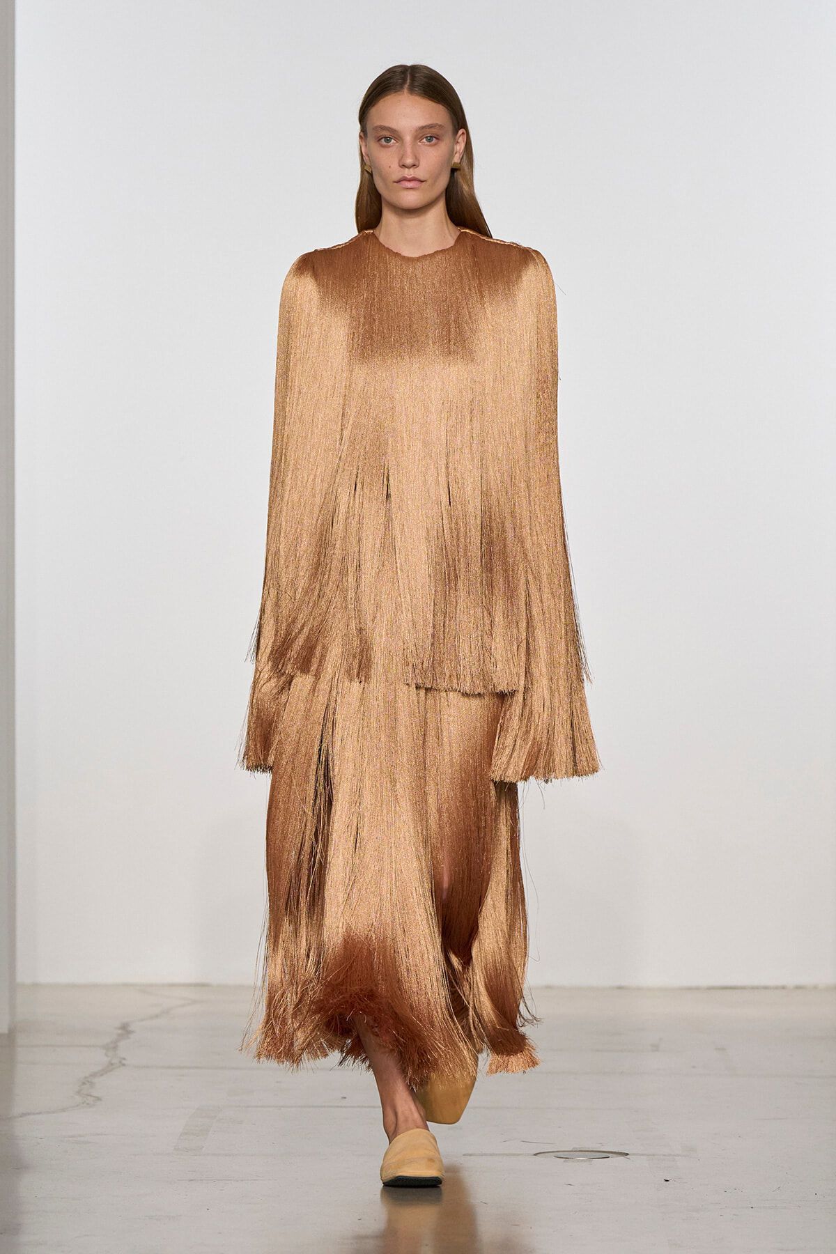 Model walking the runway in a metallic bronze fringe dress with layered, floor-length fringe panels and neutral flats