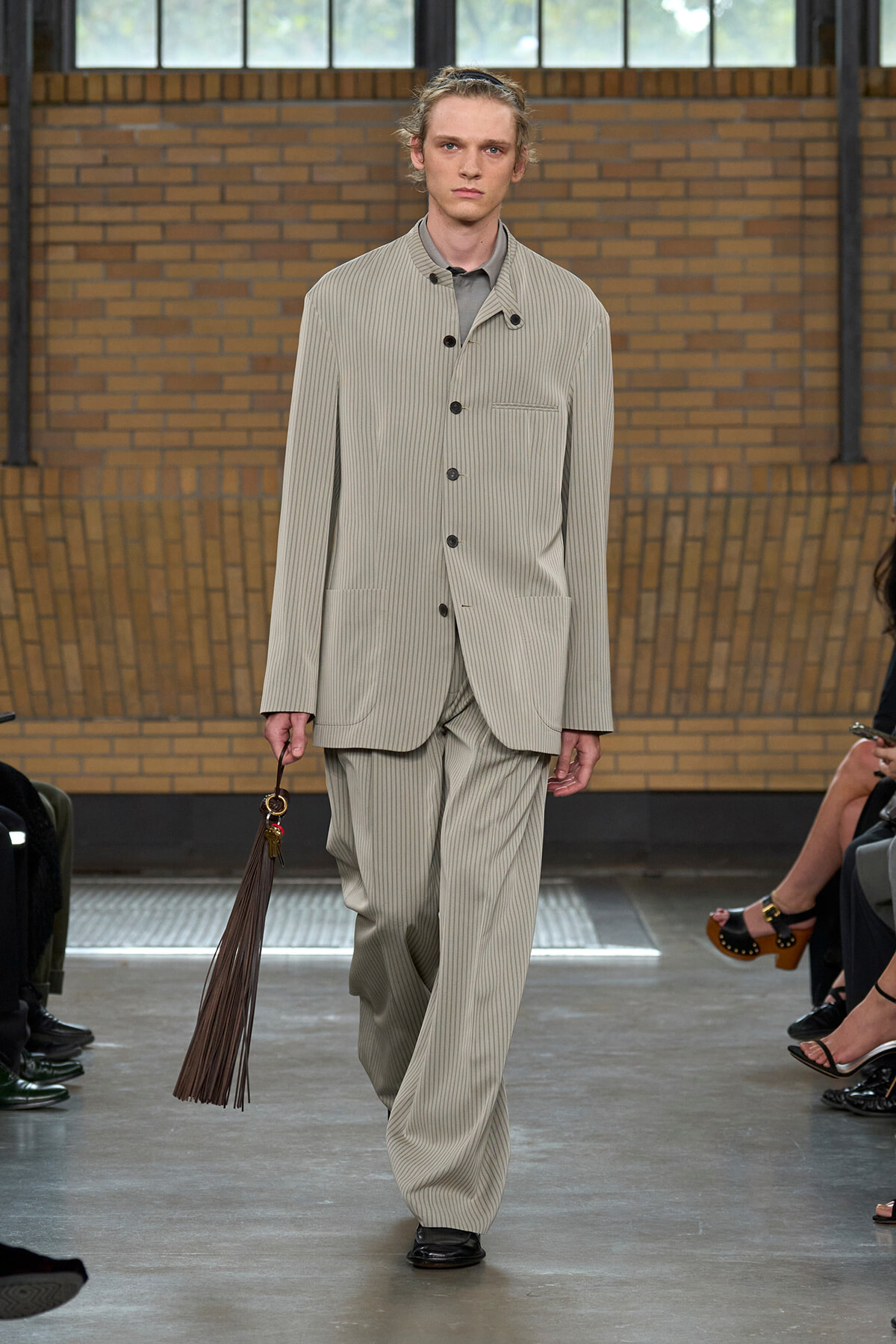 Male model wearing a light gray pinstriped oversized suit with wide pants, walking the runway with a brown fringe accessory from his hand.