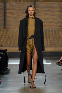 Model walks a runway in a long black coat over a gold satin romper and matching headscarf, neutral expression, brick wall backdrop.