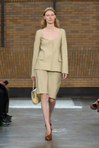 Model on a runway wearing a light beige tailored blazer and matching knee-length shorts, carrying a beige bag and brown heels.