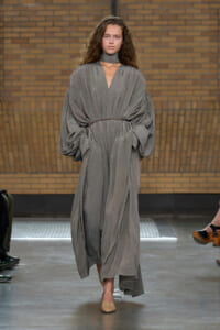 Runway model wearing a loose, gray draped dress with voluminous sleeves and a thin belt on a fashion show catwalk.