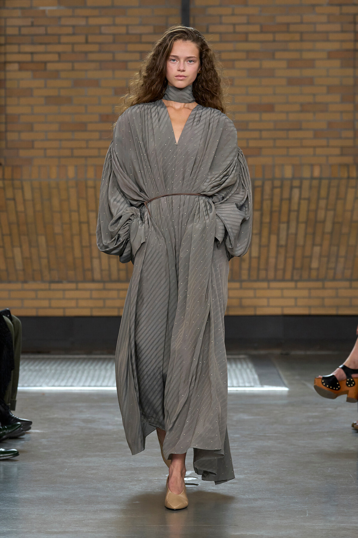 Runway model wearing a loose, gray draped dress with voluminous sleeves and a thin belt on a fashion show catwalk.