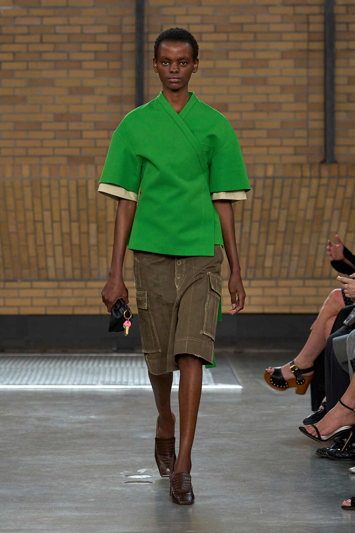 Model walks the runway wearing a bright green kimono-style top and olive cargo shorts, holding a small black clutch with keys.