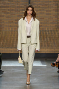 Model walks a fashion runway in a pale beige oversized blazer and matching trousers, striped shirt, brown belt, and beige heels, carrying a small beige handbag.