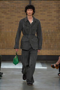 Male model walks a runway in a washed dark gray utility jacket with belt and matching wide-leg pants, holding a bright green handbag in his left hand.