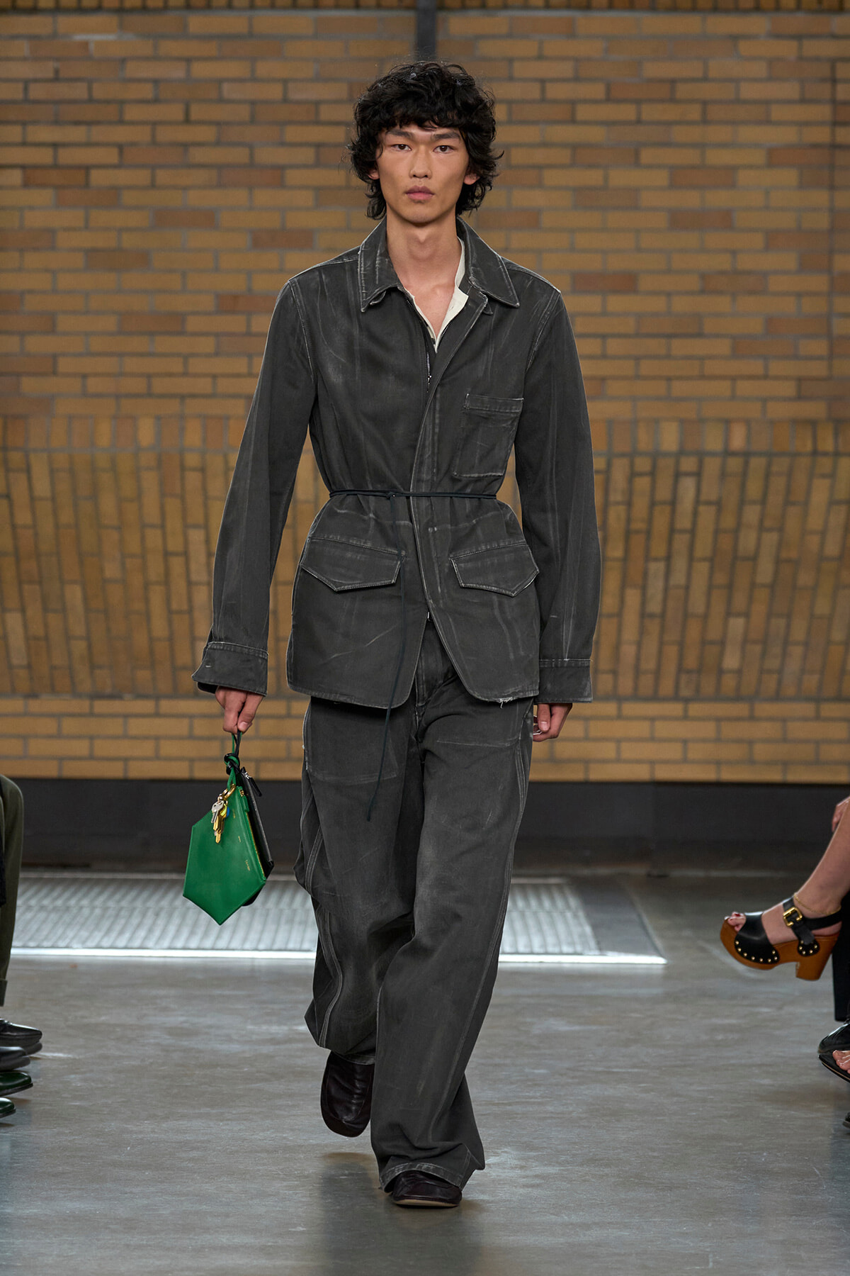 Male model walks a runway in a washed dark gray utility jacket with belt and matching wide-leg pants, holding a bright green handbag in his left hand.