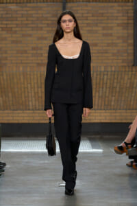 Model walks a runway in a black suit with a white bra-style top visible at the neckline, carrying a small black handbag.