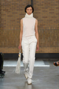 Model walking a fashion runway in a monochrome cream outfit: sleeveless turtleneck top, tailored white pants, pointed cream boots, and a long white fringe bag against a brick wall backdrop.