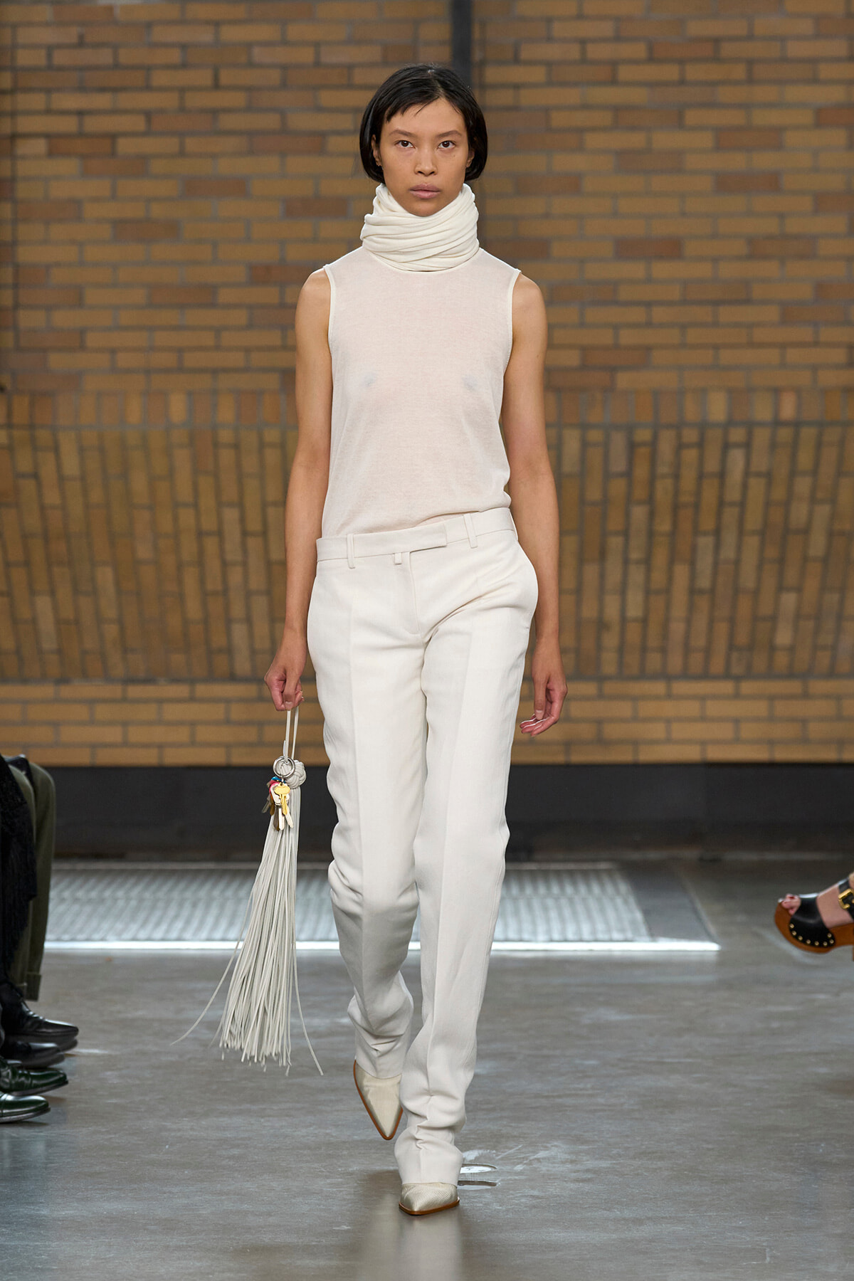 Model walking a fashion runway in a monochrome cream outfit: sleeveless turtleneck top, tailored white pants, pointed cream boots, and a long white fringe bag against a brick wall backdrop.