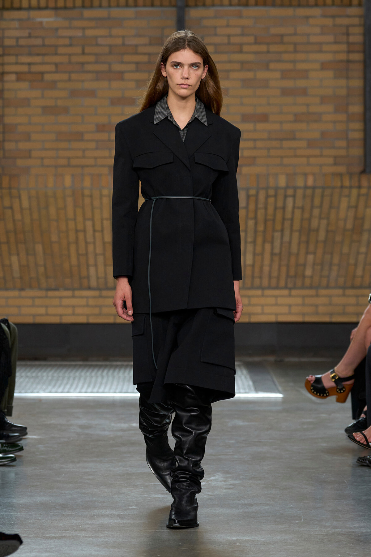 Model walking a runway in a black belted coat with high black boots, brick wall backdrop behind her.