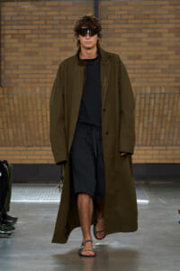 Runway model walking in a long olive-brown coat over a black outfit and sandals, wearing dark visors/sunglasses under a brick backdrop.