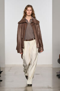 Model walking runway in a brown leather jacket, gray polo top, and cream wide-leg trousers with a brown belt.