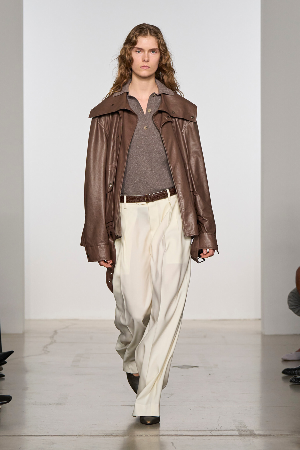 Model walking runway in a brown leather jacket, gray polo top, and cream wide-leg trousers with a brown belt.