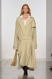 Model on a runway wearing a long beige trench coat with a belt, white collar shirt, and brown boots; holding a tan fringe accessory in her left hand.