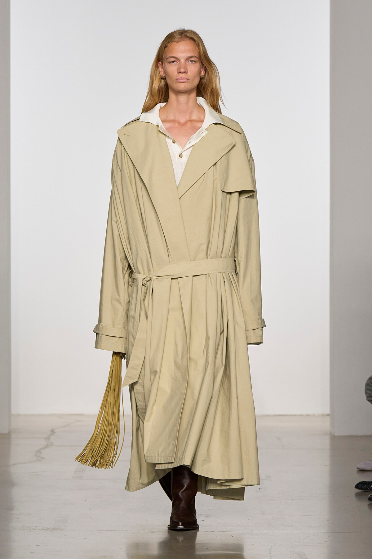 Model on a runway wearing a long beige trench coat with a belt, white collar shirt, and brown boots; holding a tan fringe accessory in her left hand.