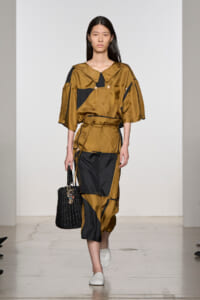 Runway model wearing a tan and black color-blocked outfit with wide sleeves and a belt, carrying a black woven handbag.