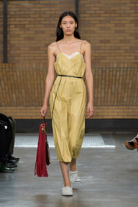 Model on a runway wearing a pale yellow sleeveless jumpsuit with a thin black belt, white top underneath, carrying a red fringe handbag.